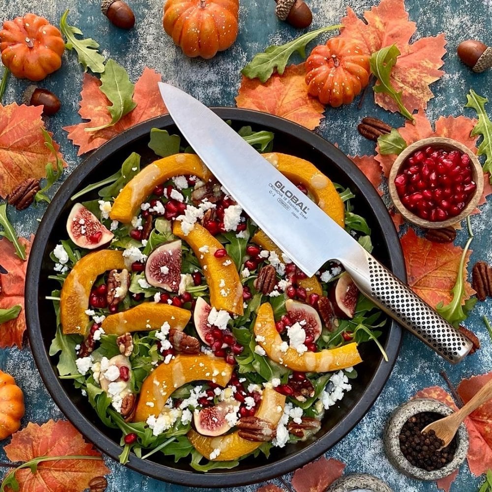Fall-themed salad with pumpkins, figs, and pomegranate seeds on a blue surface with a Global knife.