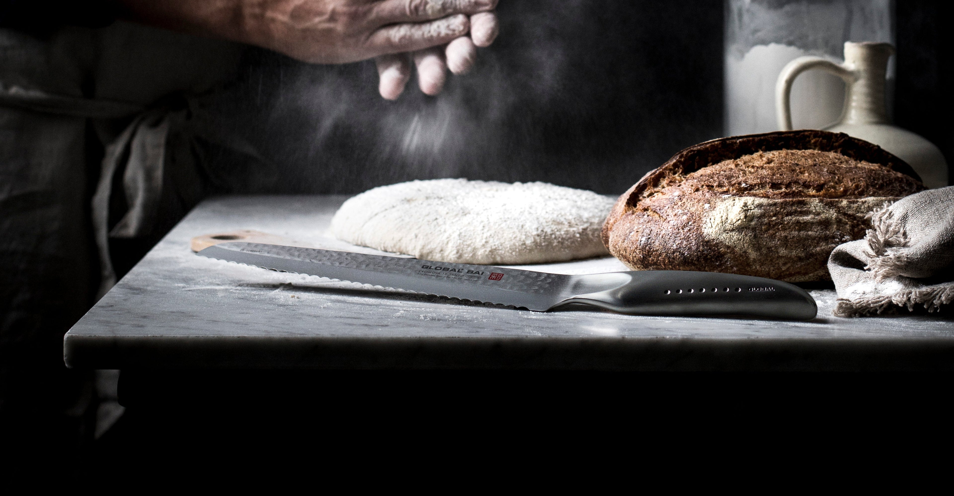 Global SAI-05 serrated bread knife next to baker baking sourdough. Serrated blade to effortlessly slice sourdough and crusty homemade breads. Baker patting flour over bread dough next to knife.