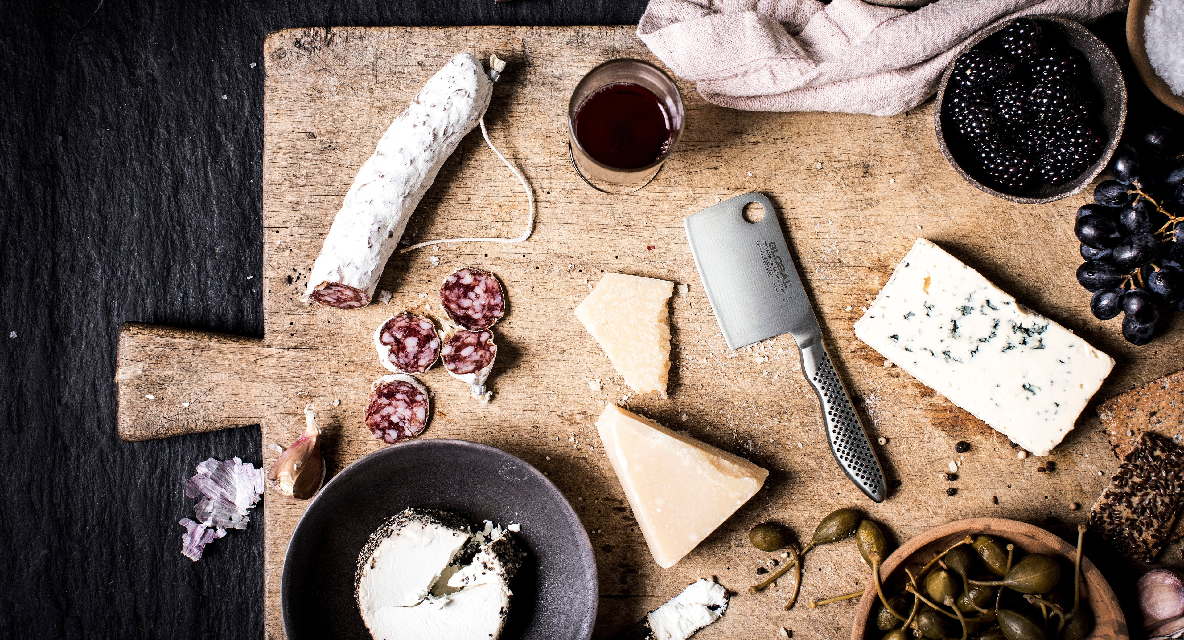 Global's small kitchen knife for chopping cheese and herbs on wooden cutting board next to charcuterie meats. Cheese board with Global's mini herb and cheese slicer. Stainless steel cheese knife.
