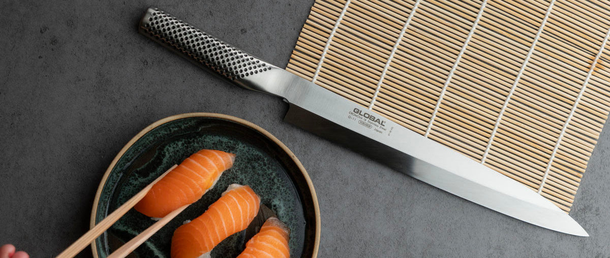 A high-angle shot of a Japanese sashimi knife resting on a bamboo mat, with a plate of salmon nigiri and chopsticks held by a hand on a dark grey surface.