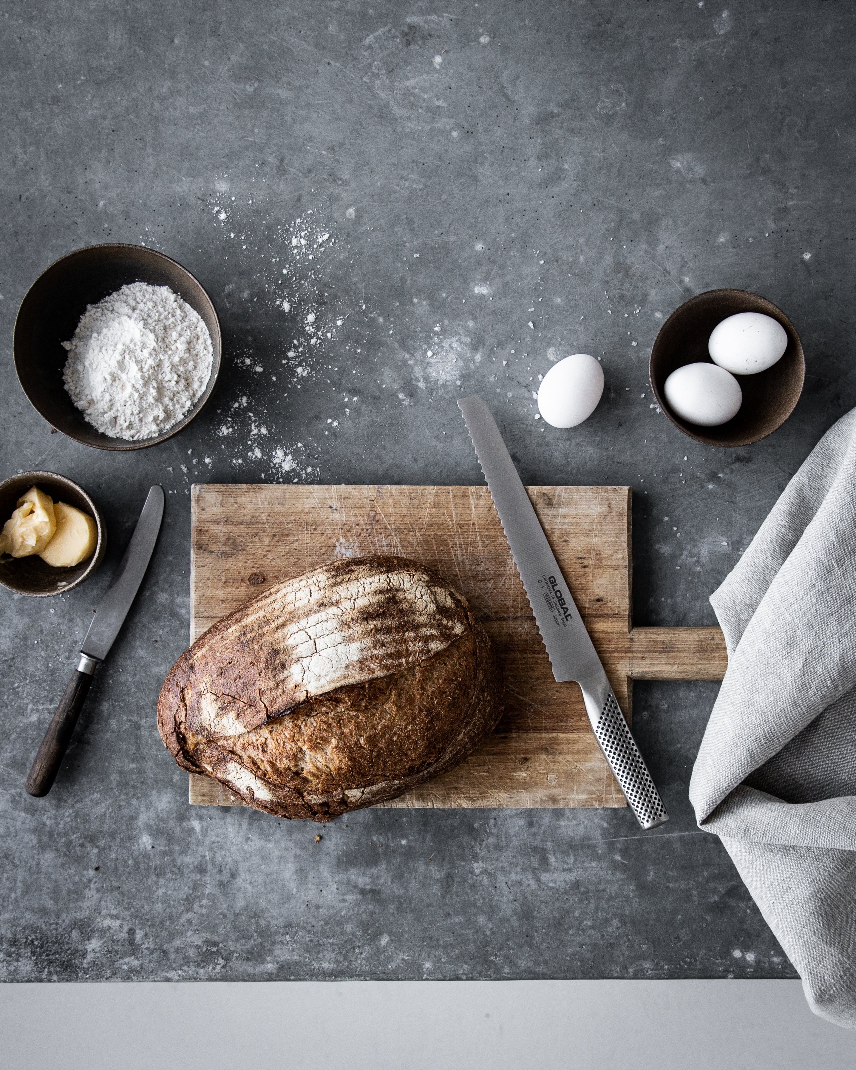 Award-wining Global G-9 serrated bread knife next to hot cross buns, jam and coffee. A stainless steel bread knife with serrated blade for effortlessly slicing crusty sourdough bread.