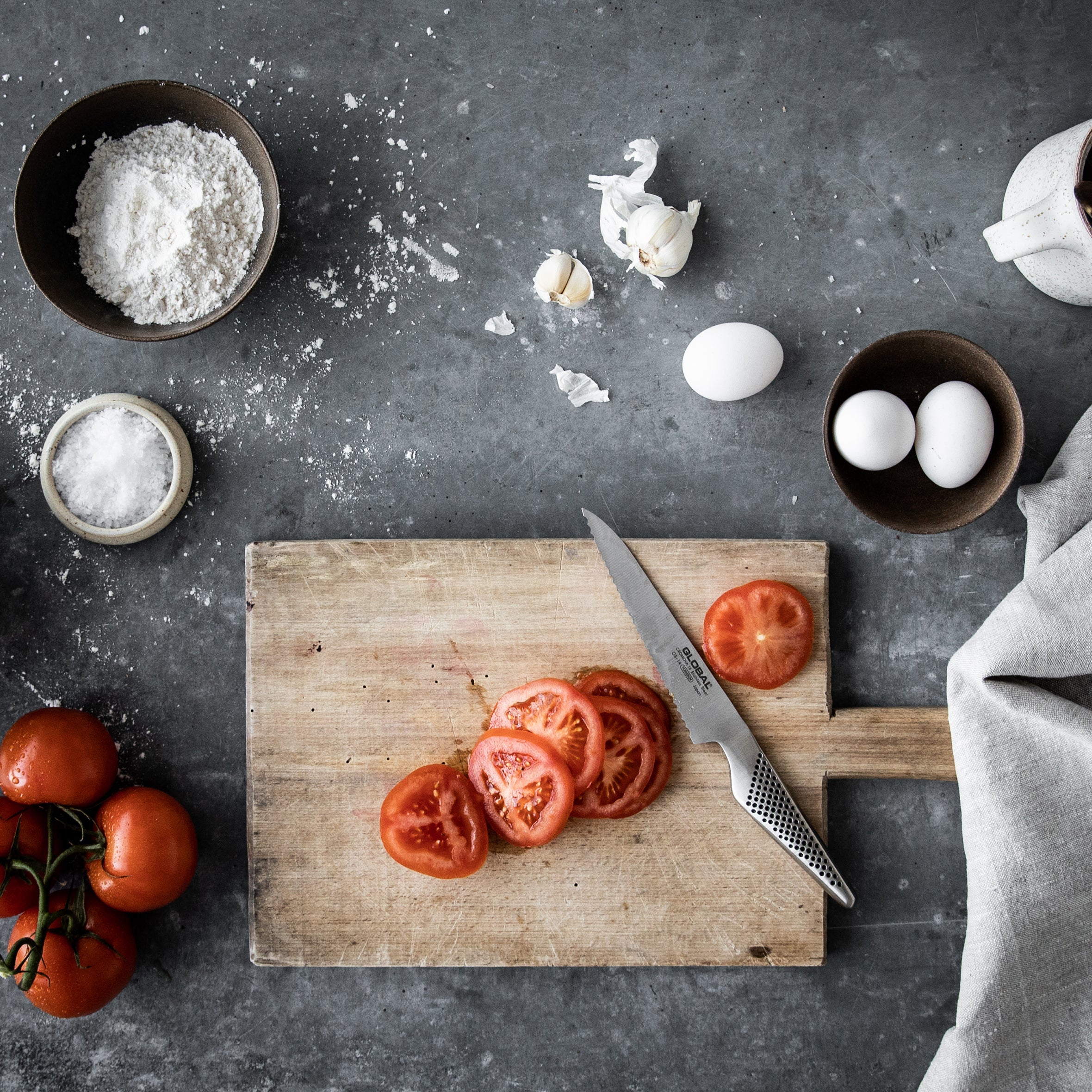 GLOBAL serrated utility knife with scalloped blade next to sliced tomatoes. Best Japanese utility knife for cutting vegetables and medium sized kitchen tasks.