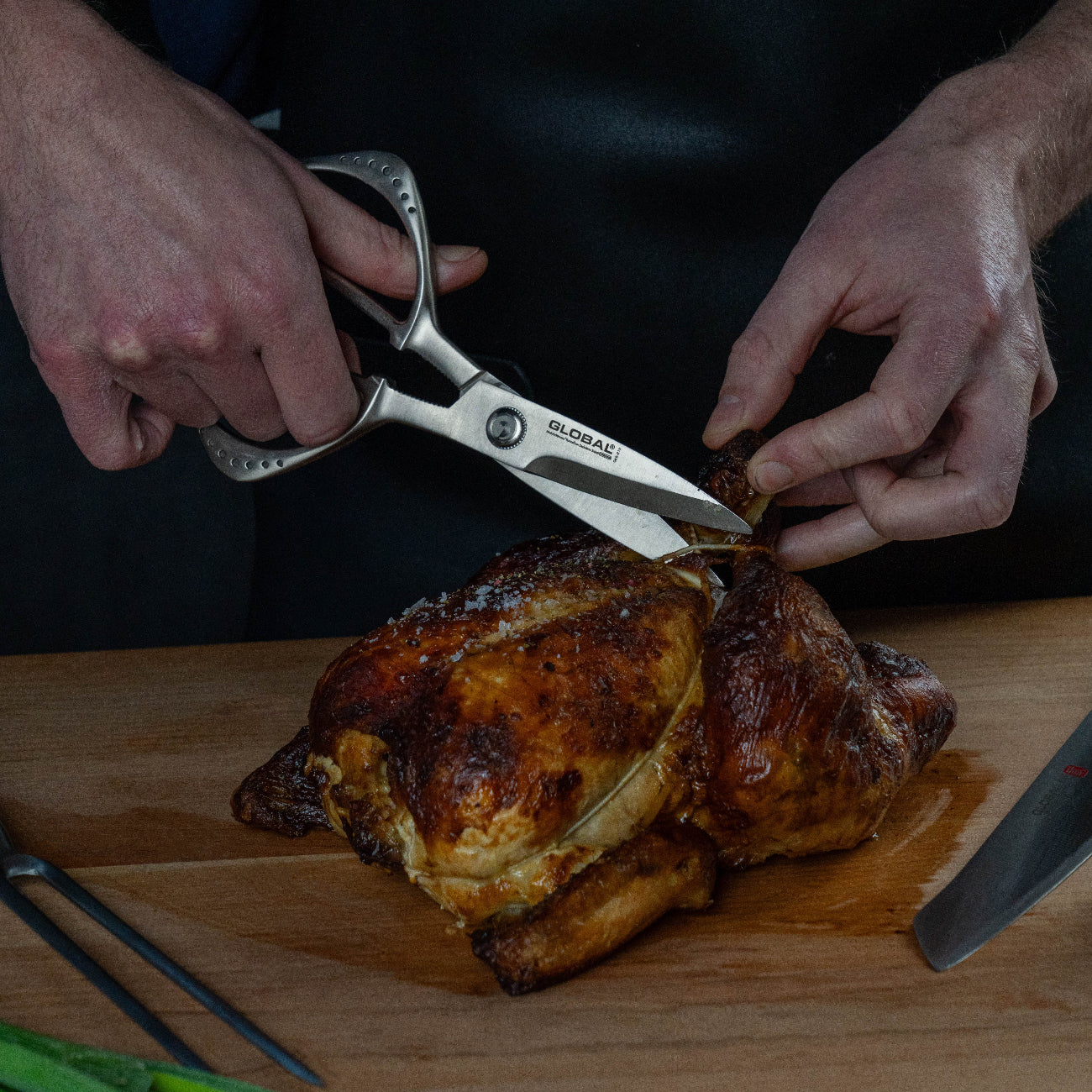 Person using kitchen shears to cut into a roasted chicken on a wooden cutting board.