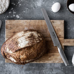 Loaf of sourdough bread on a wooden cutting board with a Global bread knife and eggs in the background. Serrated bread knife for bakers.