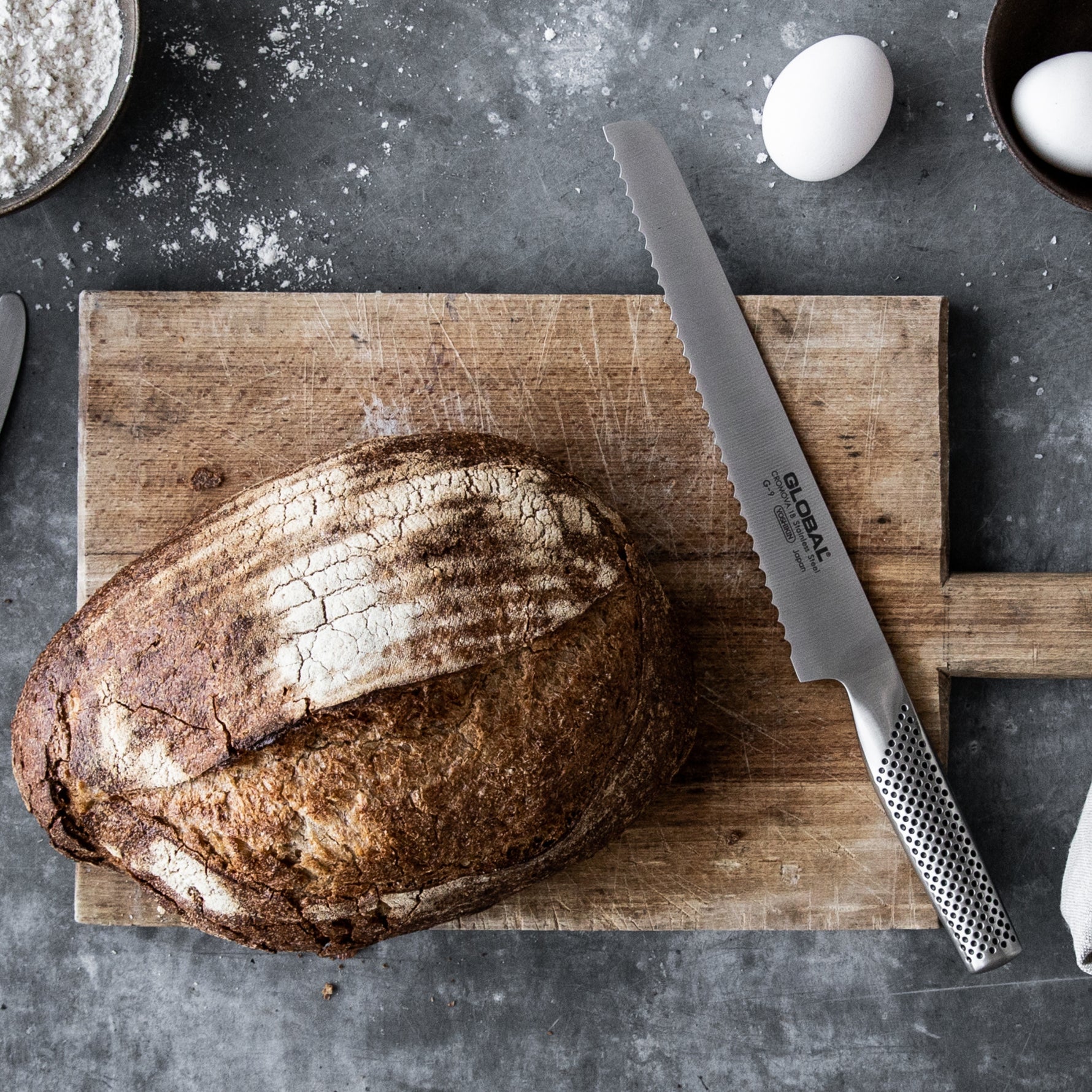 Loaf of sourdough bread on a wooden cutting board with a Global bread knife and eggs in the background. Serrated bread knife for bakers.
