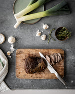 Sliced roast meat on a wooden cutting board with Global G-3 Meat Carcing Knife, surrounded by leeks, garlic, and herbs on a gray surface.