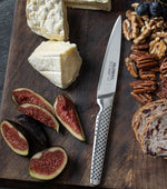 Wooden cutting board with cheese, figs, nuts, and bread, featuring a small kitchen knife, the Global GSF-22 stainless steel utility knife.