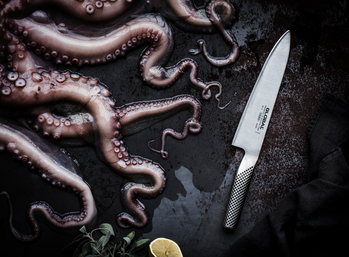 Stainless steel kitchen knife next to a cut octopus on a dark surface. Global Japanese chef's knife, the G-2 all stainless steel cook's knife.