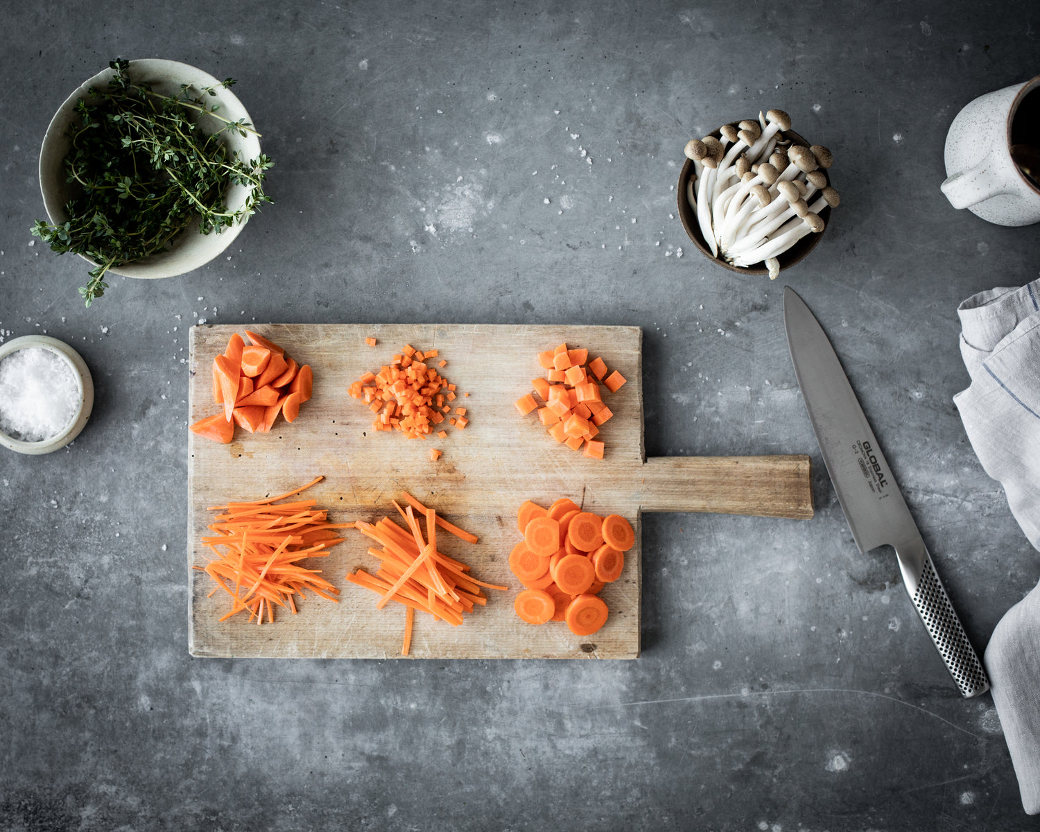 Wooden cutting board with sliced, diced, chopped and julienned carrots, a Global G-2 Chef's knife, and ingredients on a grey surface. Ultra sharp stainless steel all-purpose kitchen knife.