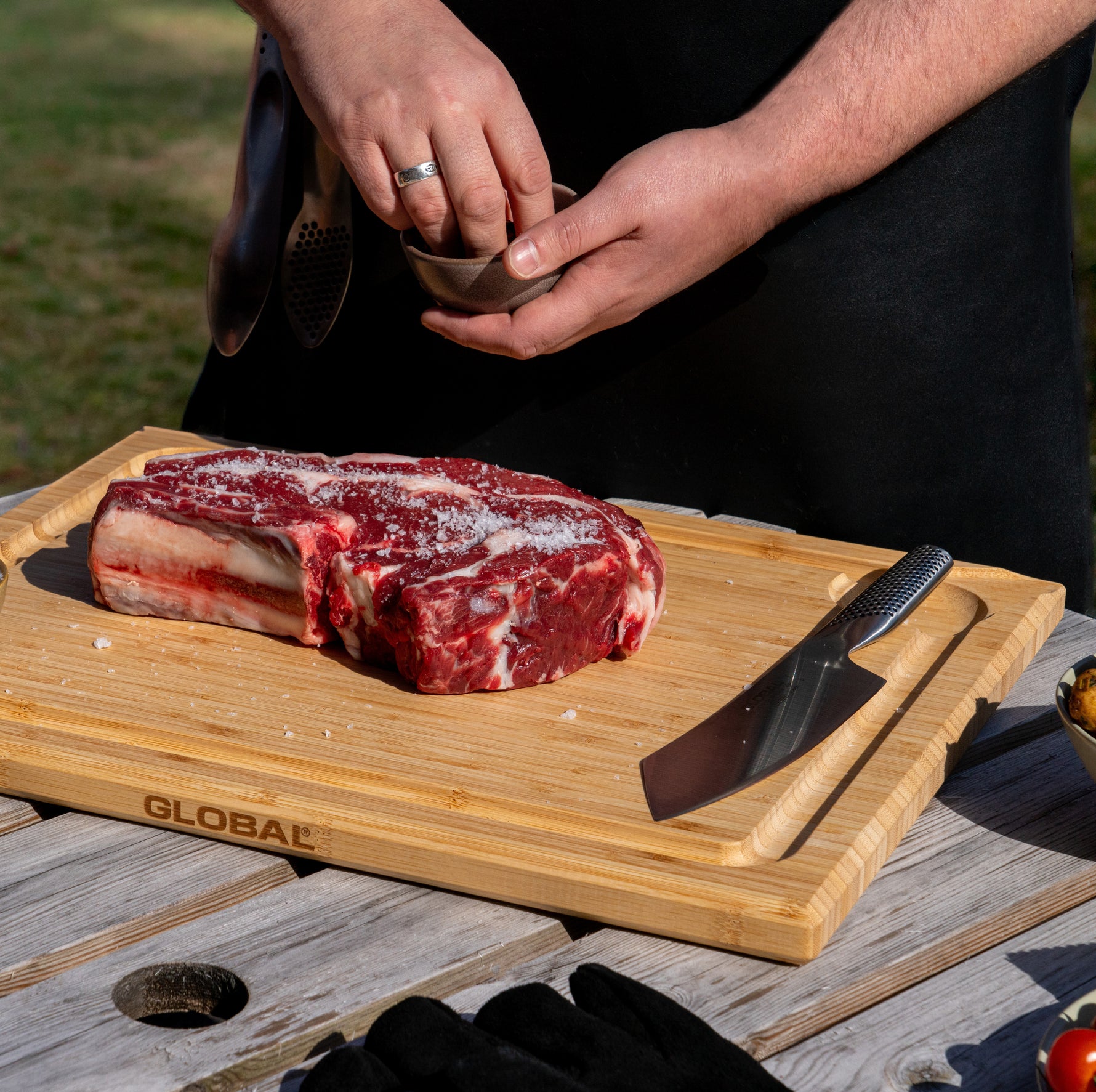 Raw steak on Global bamboo wooden cutting board with a Global Japanese bbq knife and black leather bbq gloves being used by person cooking outdoors.