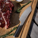 Raw steak with seasoning, a knife, and rosemary on a wooden cutting board with the Global Japanese BBQ knife.