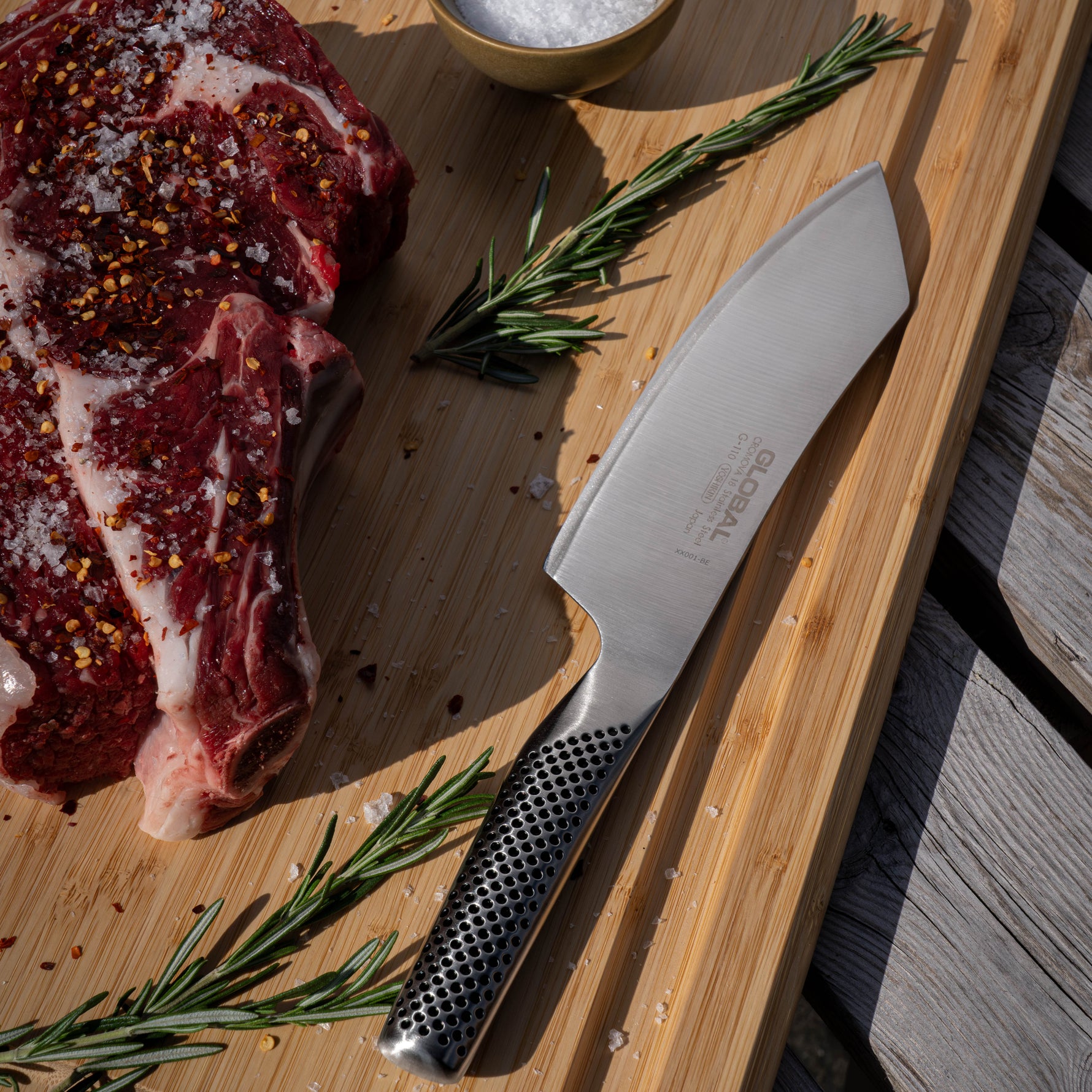 Raw steak with seasoning, a knife, and rosemary on a wooden cutting board with the Global Japanese BBQ knife.