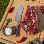 Raw steak on a wooden cutting board with the Global barbecue knife, salt, and red peppers.