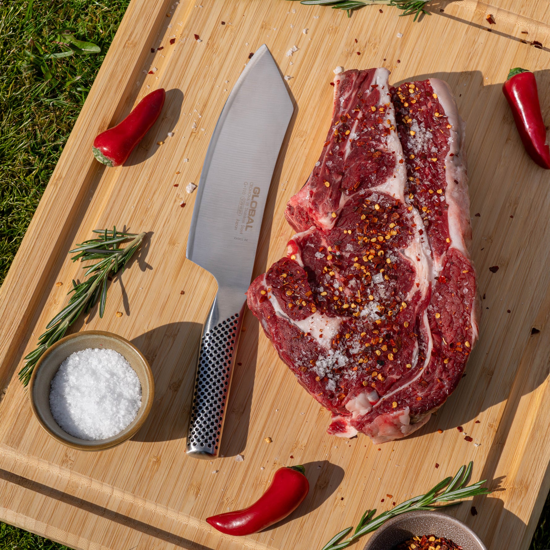 Raw steak on a wooden cutting board with the Global barbecue knife, salt, and red peppers.