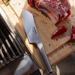 Global stainless steel barbecue chef's knife on a wooden cutting board with raw meat and peppers, next to a grill.