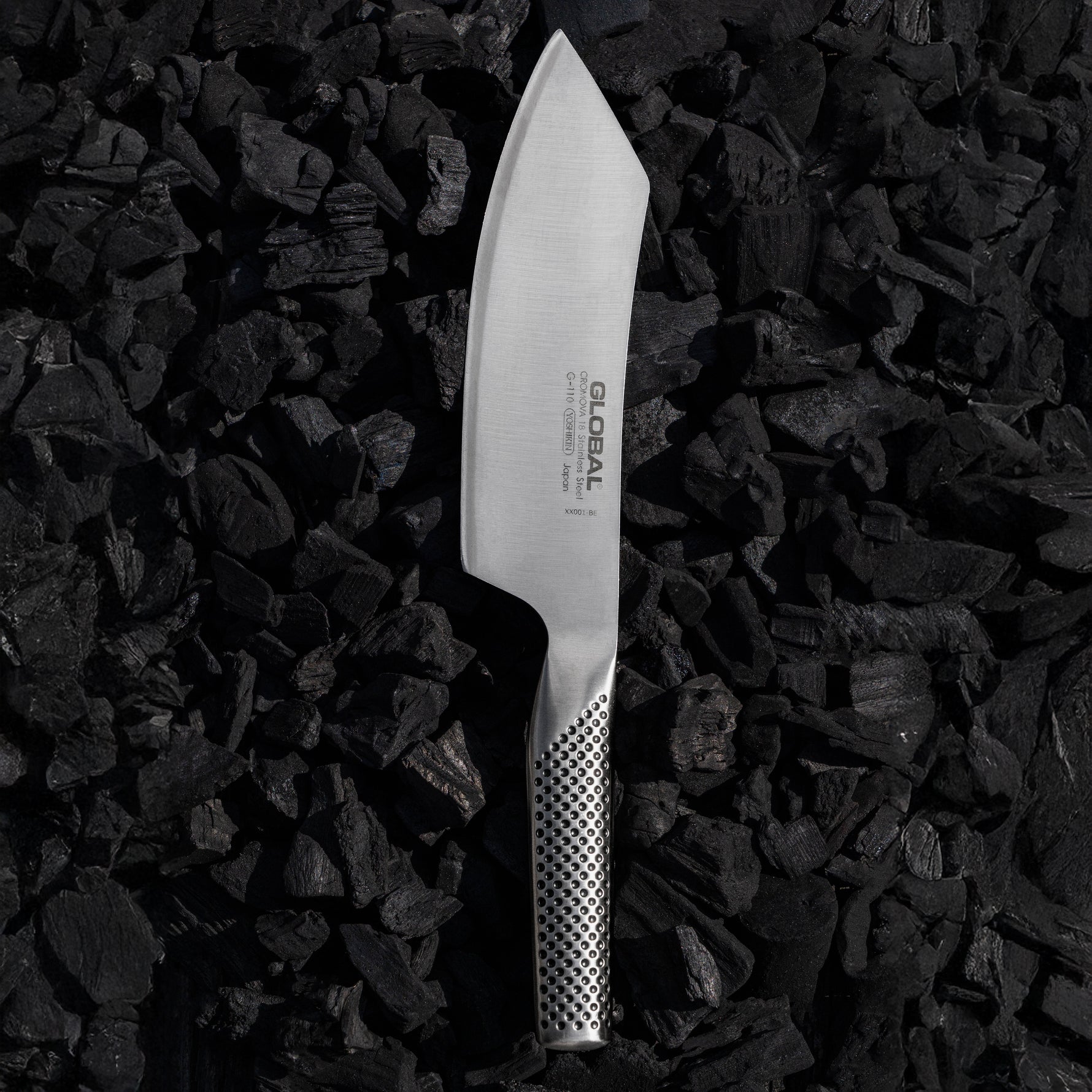 Japanese BBQ Knife by Global on charcoal. Stainless steel barbecue knife with large, curved blade and pointed tip.