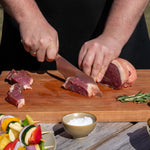 Outdoor cook cutting raw meat on a wooden board using Global Japanese BBQ chef's knife with vegetables and salt in the background