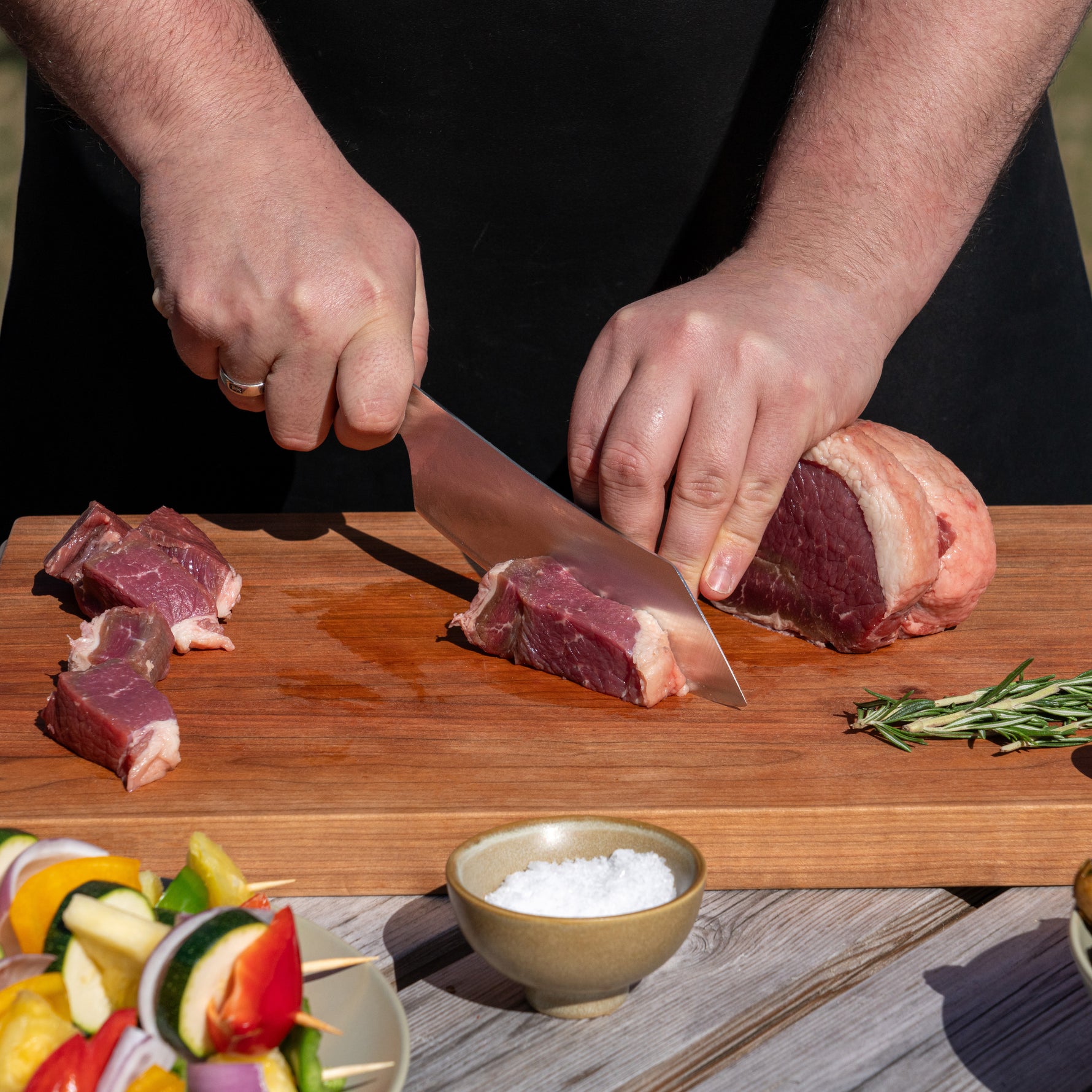 Outdoor cook cutting raw meat on a wooden board using Global Japanese BBQ chef's knife with vegetables and salt in the background