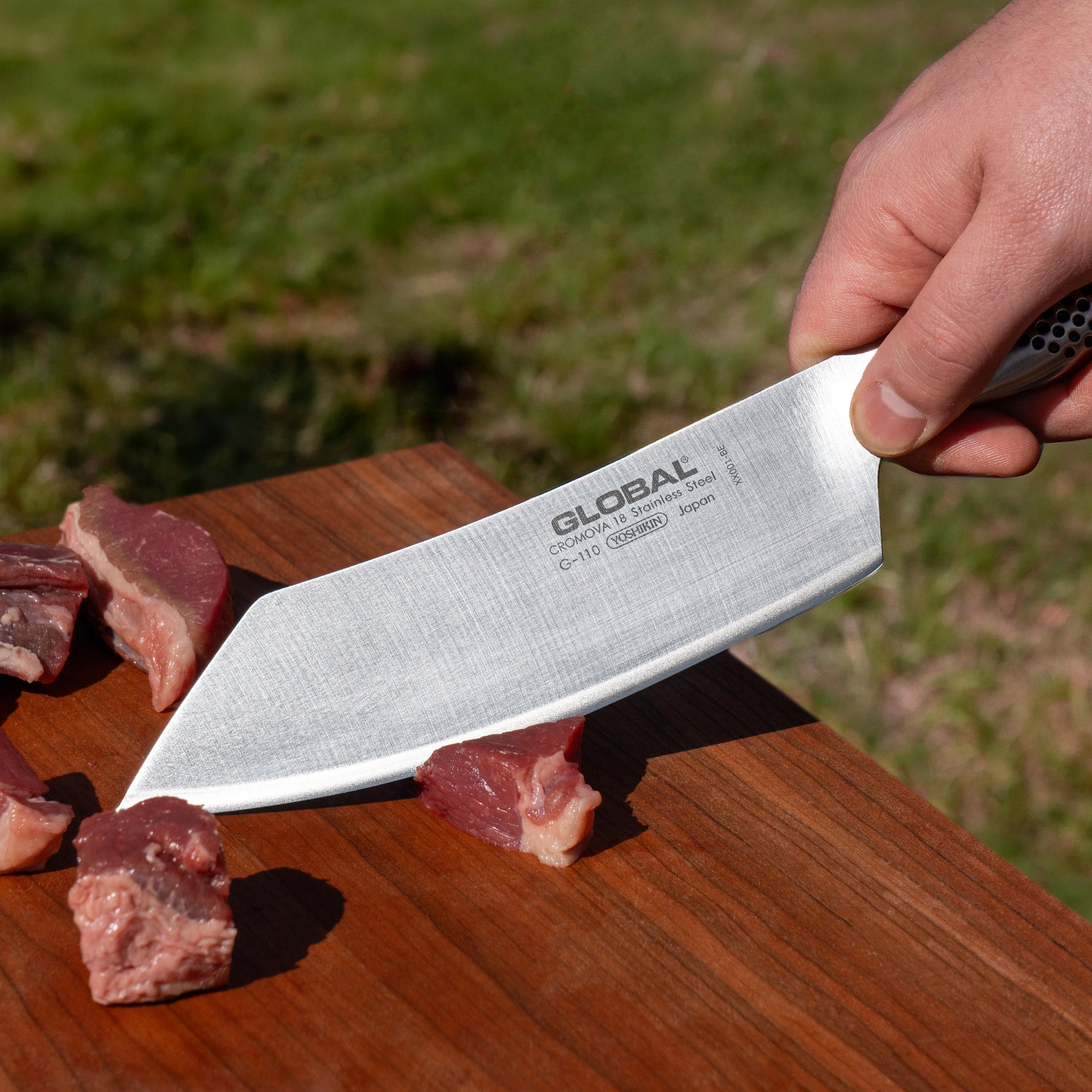 Chef holding a Global brand BBQ knife over chopped meat on a wooden cutting board with a grassy background.