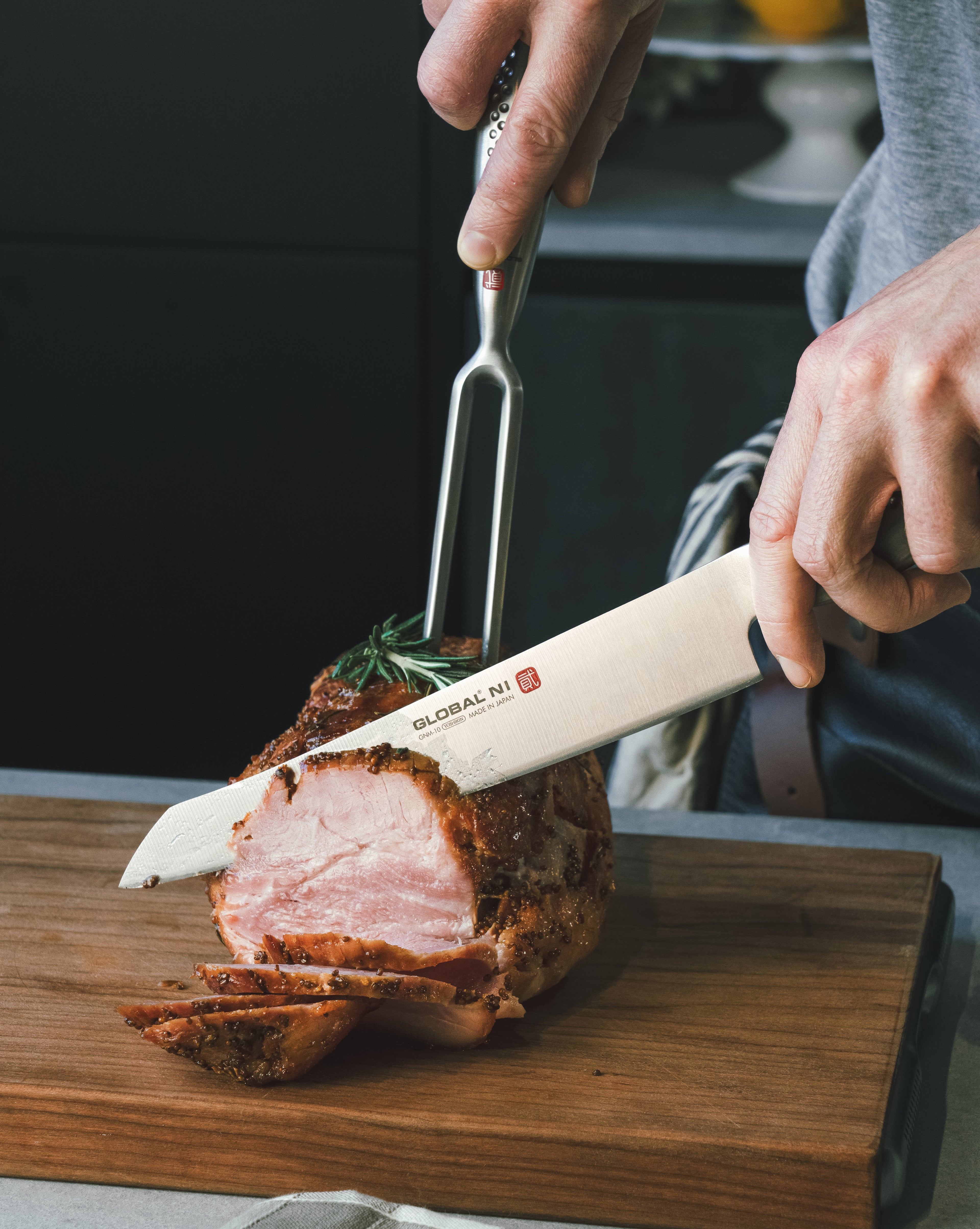 Home cook cutting into a roasted meat dish with a Global Ni Carving knife and fork on a wooden board. Japanese stainless steel carving knife and fork set.
