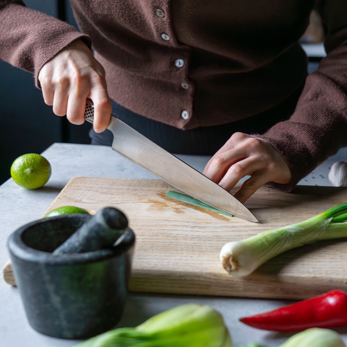 Home cook using ultra sharp Chef's Knife, the Global G-2 Cook's knife, for slicing onions, lime, chilli pepper and fennel on wooden chopping board.