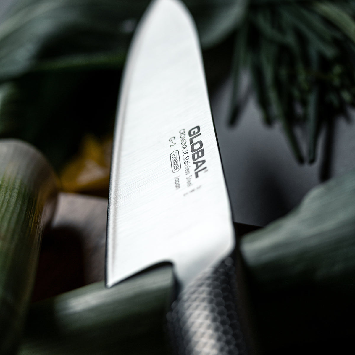 Close-up of a Global G-2 Cook's Knife ultra sharp blade with vegetables in the background for effortlessly cutting vegetables.