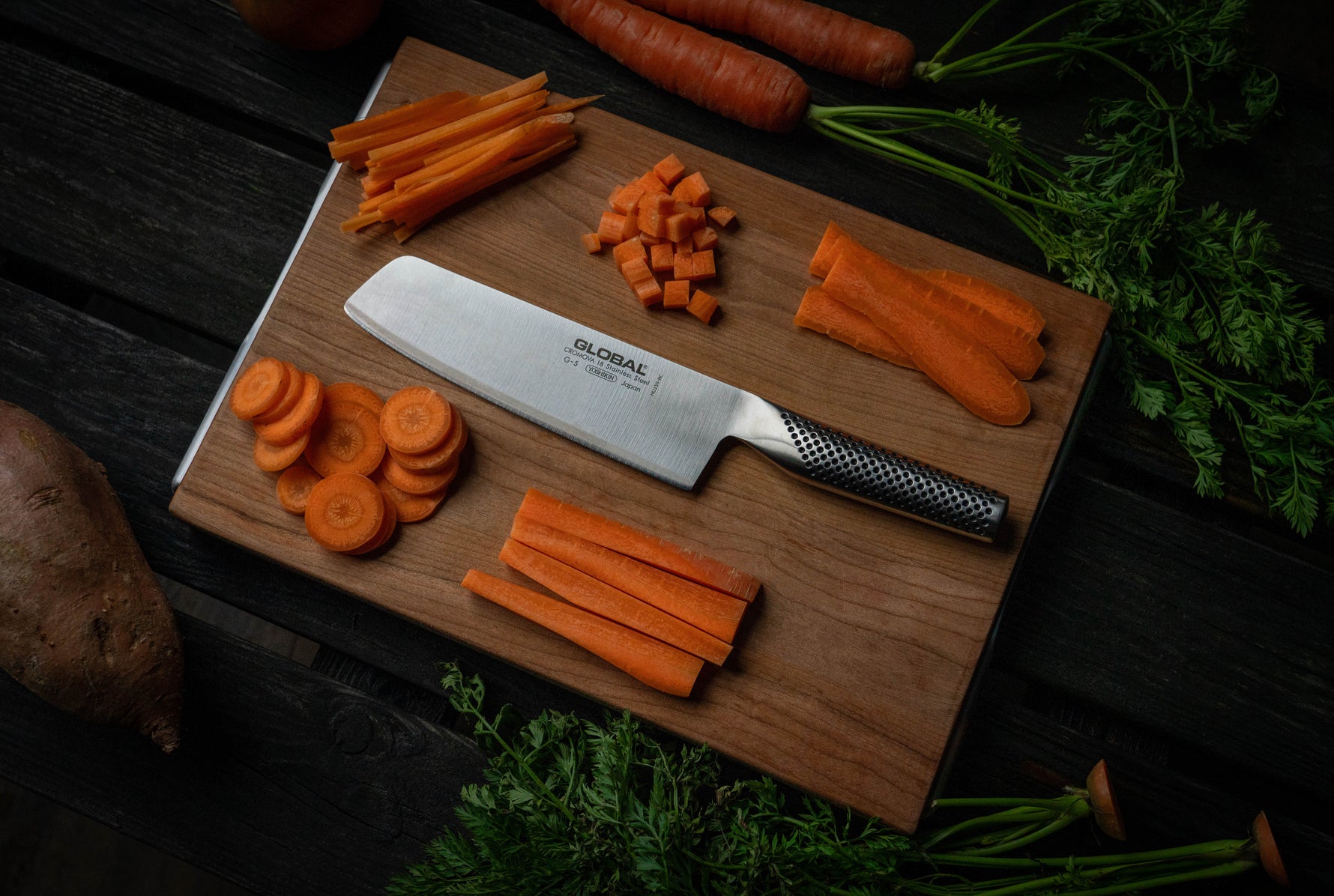 Wooden cutting board with sliced carrots and a Global kitchen knife for chopping vegetables, surrounded by fresh vegetables on a dark surface.