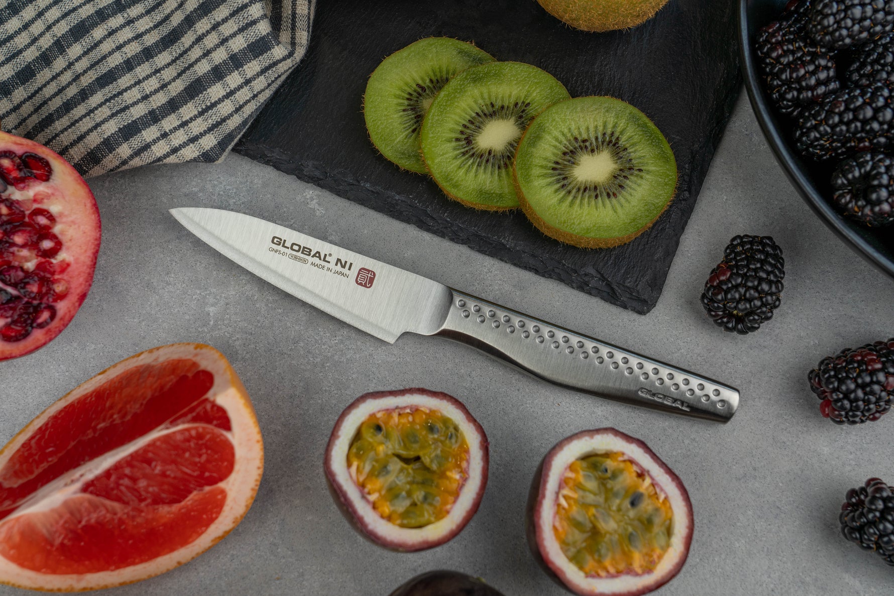 Fruits including kiwis, pomegranate, and grapefruit with a Global paring knife on a stone surface.