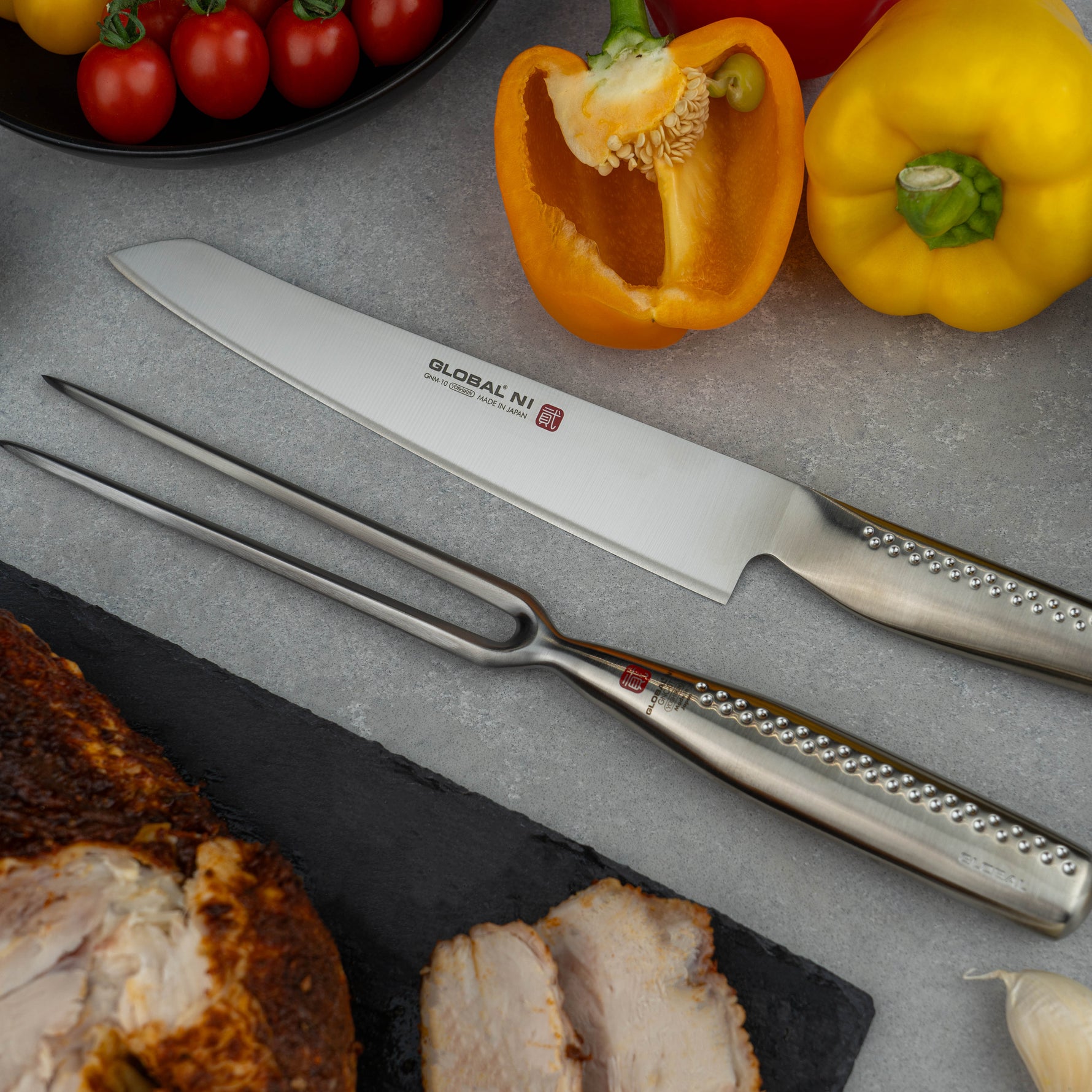 Global Japanese carving Knife and carving fork set on a grey surface with vegetables and meat in the background. Stainless steel carving set for meat, poultry, turkey, ham, roasts and more.