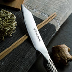 Ultra sharp Global Kritsuke knife with  slightly curved blade, pointed tip and 'Global 40th Anniversary' branding on a dark surface with noodles and chopsticks.