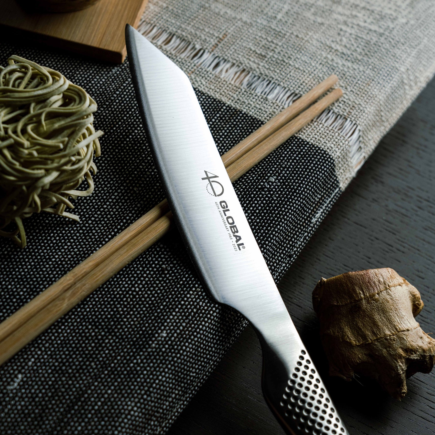 Ultra sharp Global Kritsuke knife with  slightly curved blade, pointed tip and 'Global 40th Anniversary' branding on a dark surface with noodles and chopsticks.