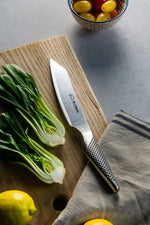 Global Kiritsuke, a Japanese Chef's Knife on a wooden cutting board with vegetables and a towel on a grey surface