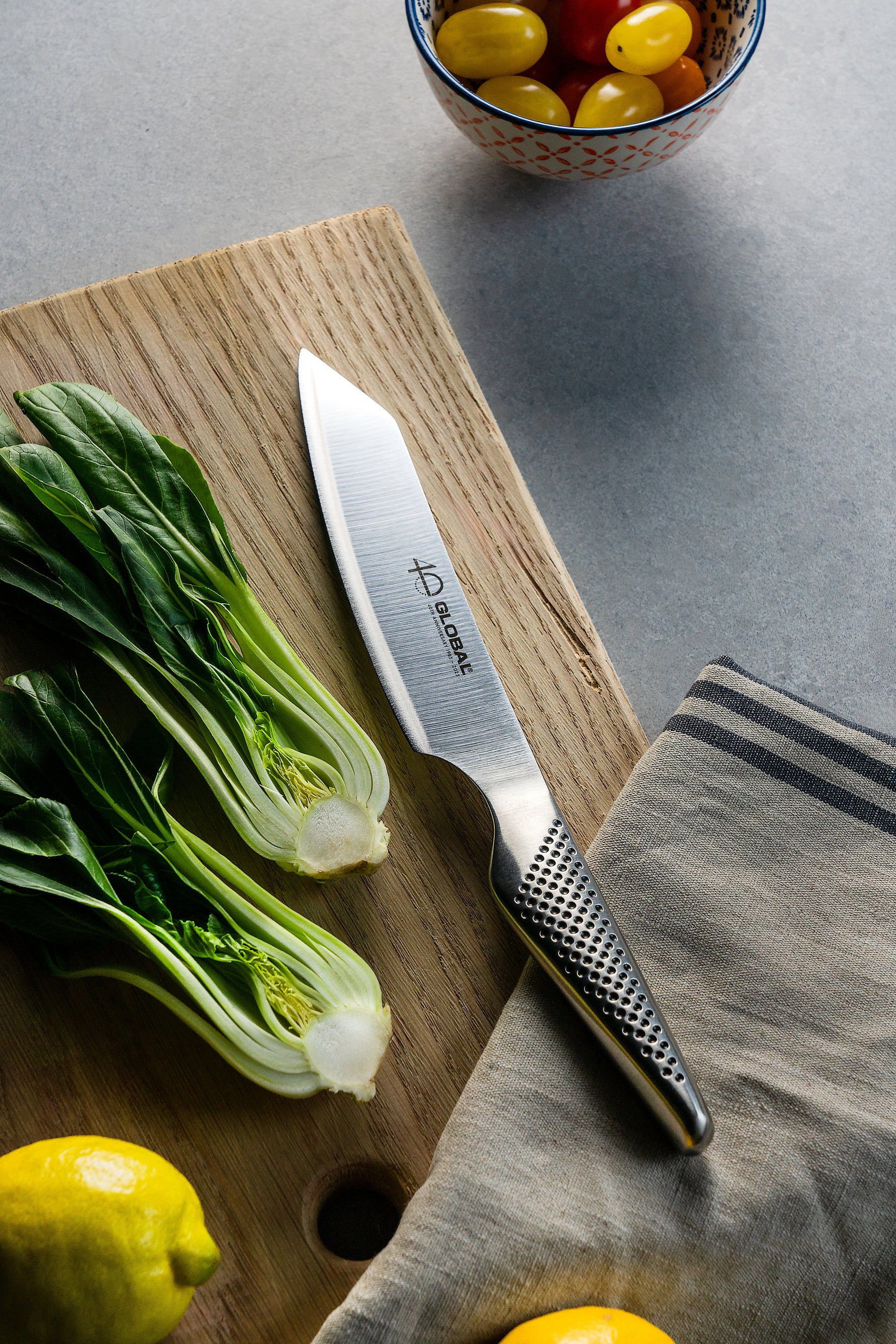 Global Kiritsuke, a Japanese Chef's Knife on a wooden cutting board with vegetables and a towel on a grey surface