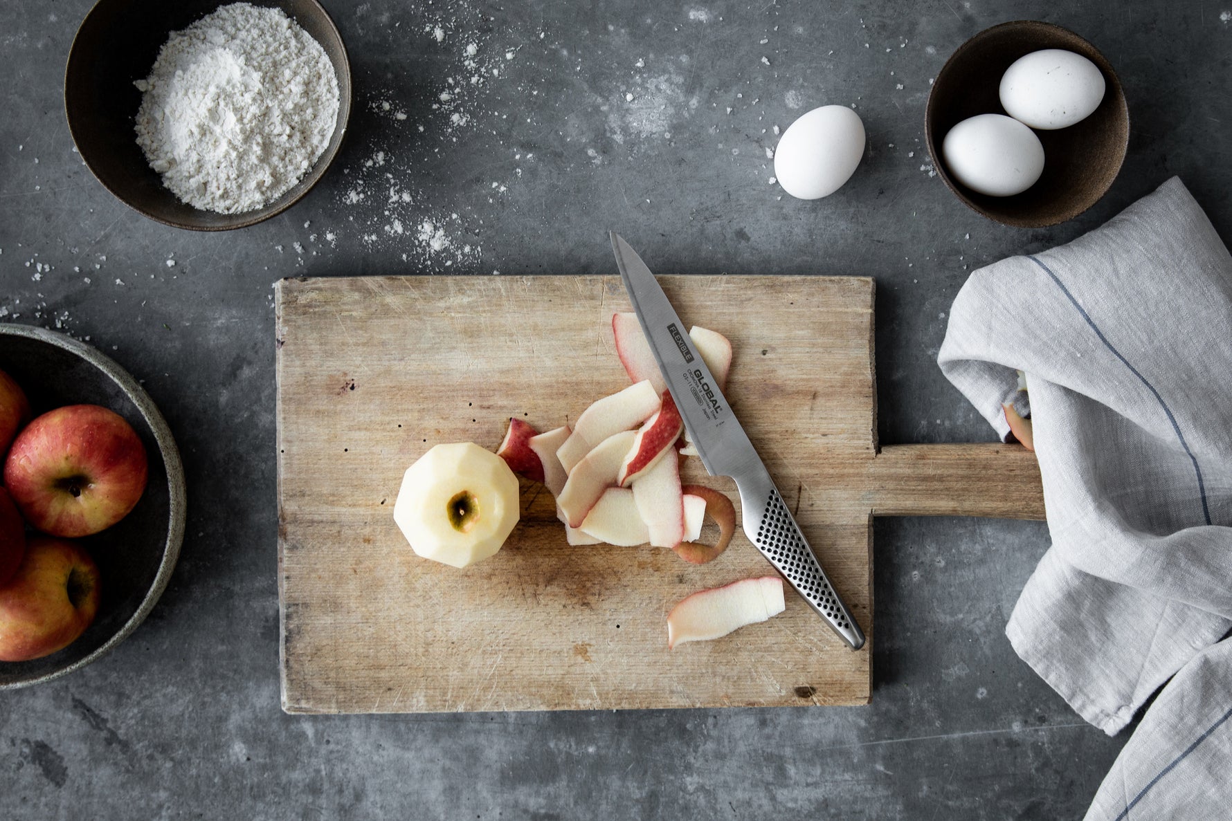 Wooden cutting board with sliced apples and a the GS-11 Flexible Utility Knife on a gray surface with bowls of flour, eggs, and more apples.