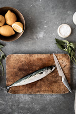 Fish on a wooden cutting board with a Global flexible utility knife, lemons, and herbs on a gray surface. The GS-11 stainless steel utility knife for cooking.