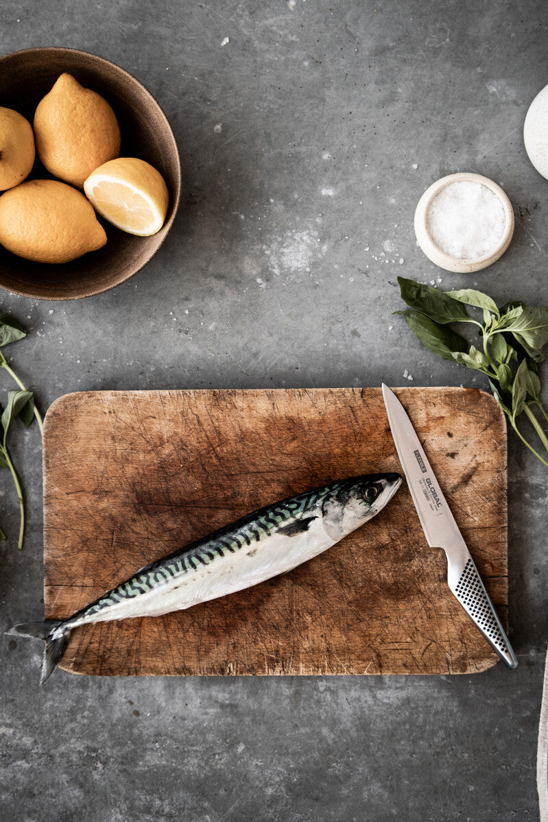 Fish on a wooden cutting board with a Global utility knife, lemons, and herbs on a grey surface. Versatile small kitchen knife for food prep.