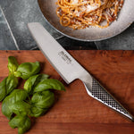 Chef's vegetable knife on a wooden cutting board with basil and pasta in the background