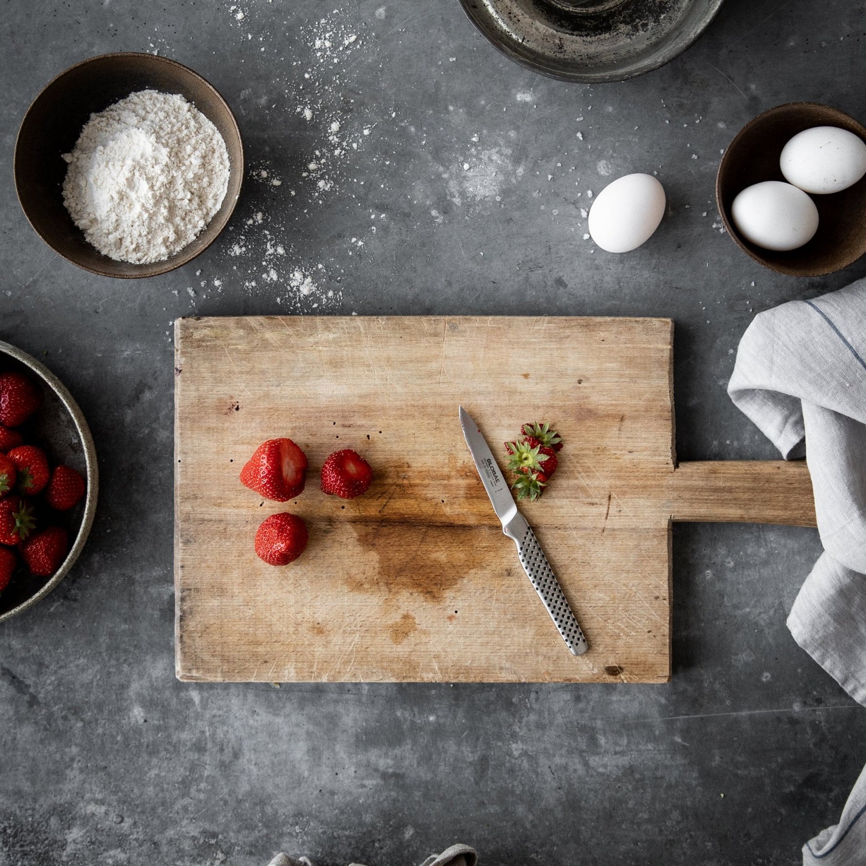 Wooden cutting board with strawberries and a Global paring knife on a gray surface with flour and eggs.