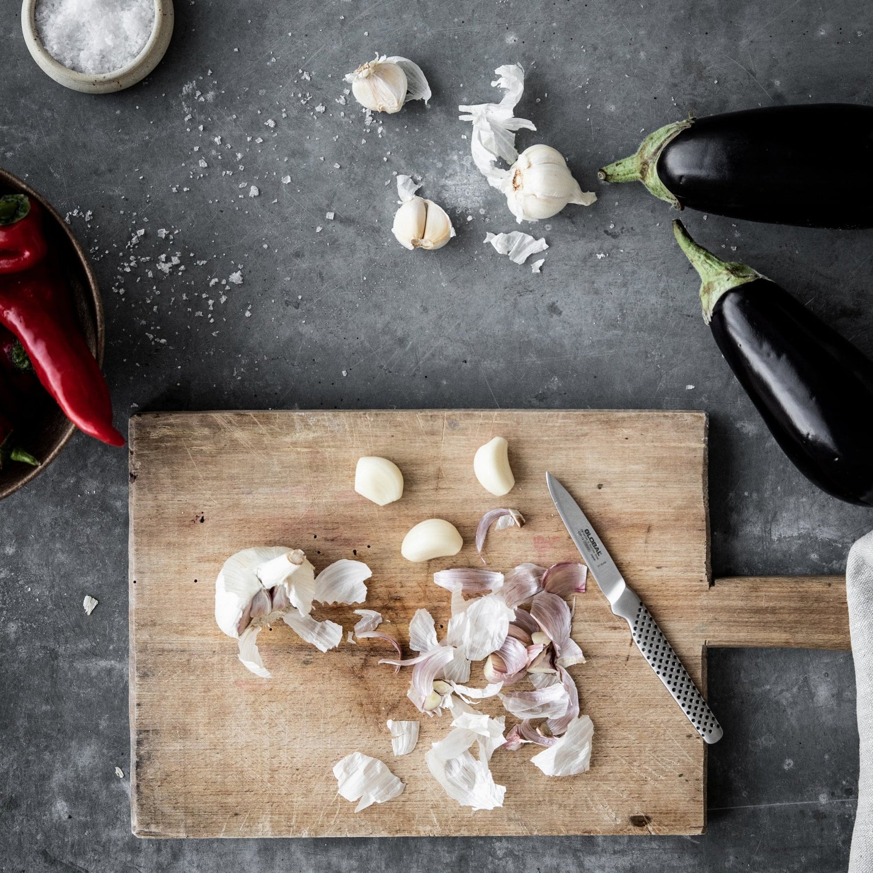 Wooden cutting board with chopped garlic and shallots, eggplant, and a Global paring knife on a dark surface, the GSF-15