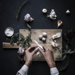 Home cook preparing garlic on a wooden cutting board with herbs and a dark background using Global GSF- Peeling knife, a small stainless steel kitchen knife.