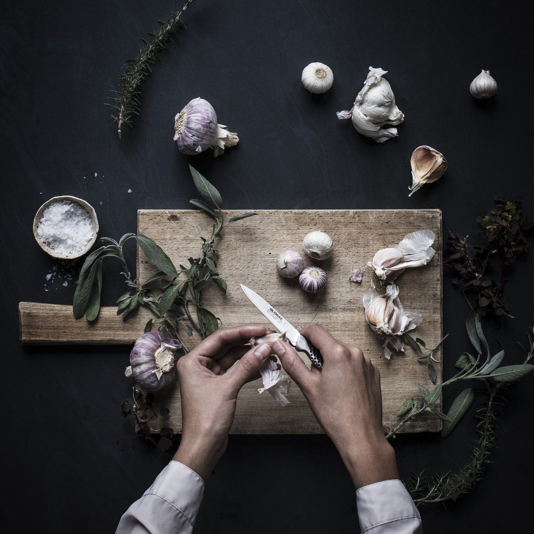 Home cook preparing garlic on a wooden cutting board with herbs and a dark background using Global GSF- Peeling knife, a small stainless steel kitchen knife.