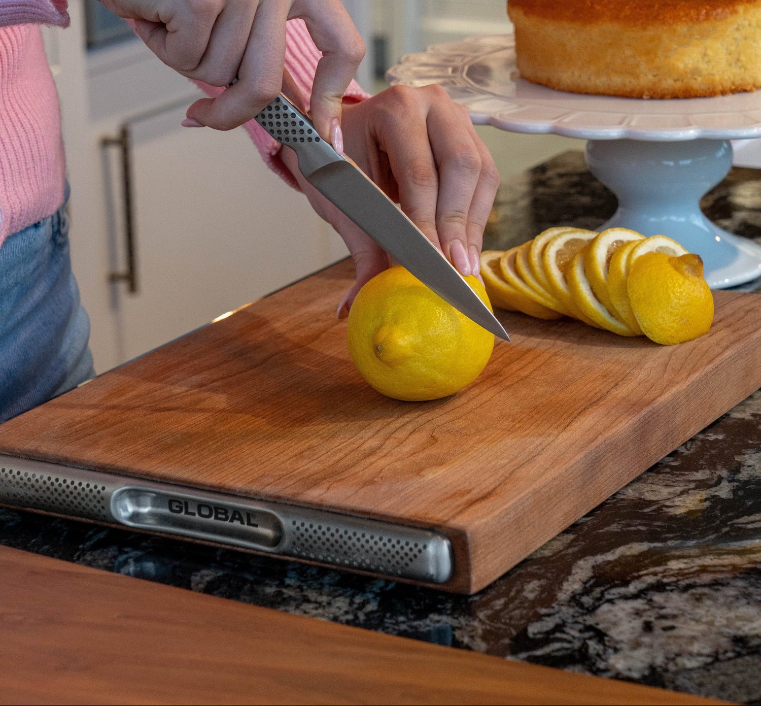 Home cook cutting a lemon on a wooden cutting board, the Global cherry wo with a cake in the background using Global utility knife, the GSF-22 small kitchen knifed chopping board,