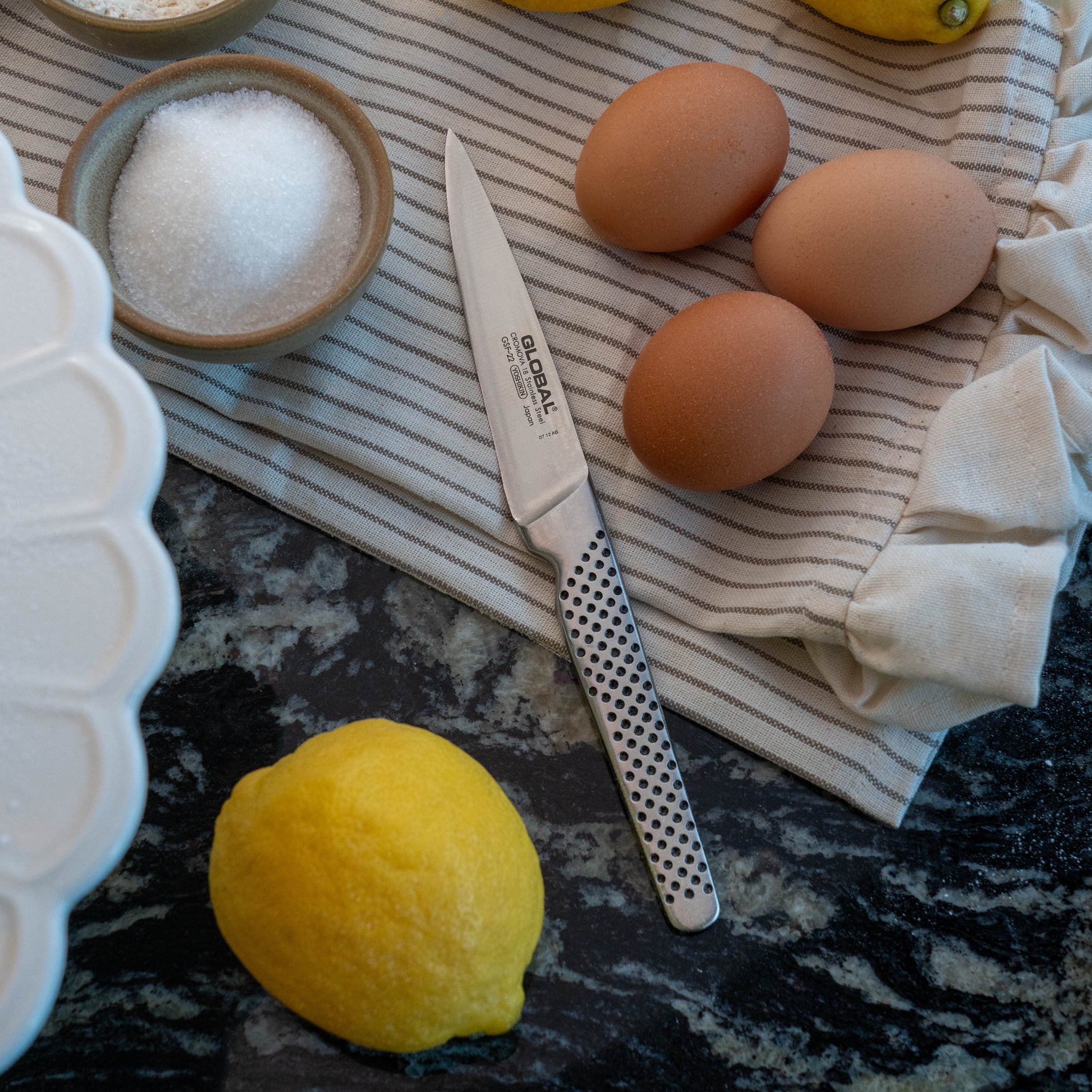Global stainless steel utility knife, eggs, lemon, and salt on a striped cloth with a dark surface. Utility knife can be used as paring knife,  peeling knife, a versatile kitchen knife.