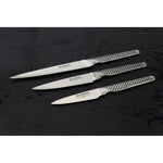 Three Global GSF small kitchen knives on a dark textured surface