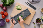 Global's adjustable mandolin slicer for cutting cucumber into precise, consistent slices on wooden chopping board next to interchangeable blades for julienning, shredding and slicing.