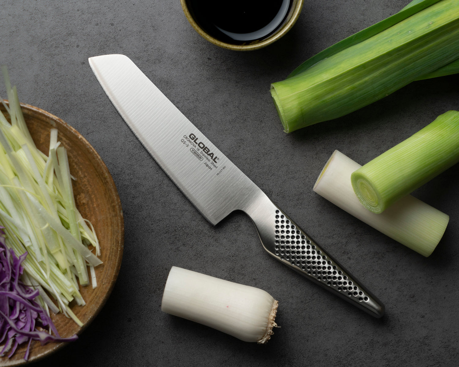 Global vegetable chopping knife on a dark surface with vegetables and a bowl of sauce.