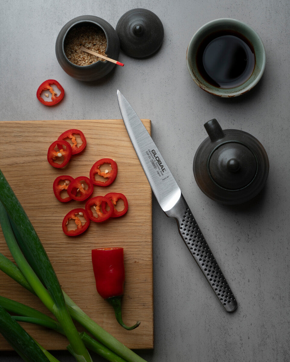 Global utility knife on a wooden cutting board with sliced red peppers, green onions, and tea-making items on a gray surface. GSH-22 Japanese stainless steel utility knife with 11cm blade, made in Japan.