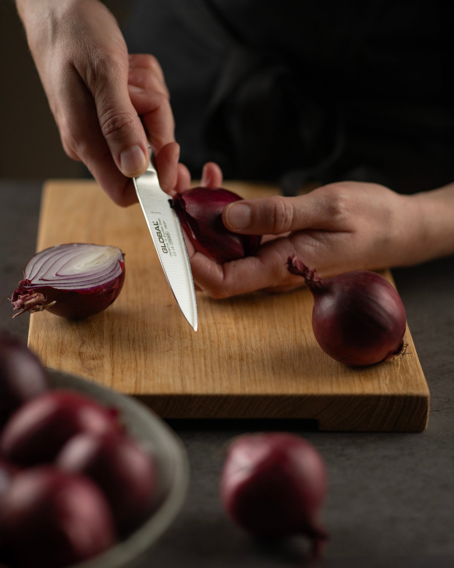 Chef cutting red onions on a wooden board with a Global peeling knife, surrounded by more onions.
