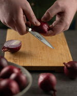 Cook cutting an onion with a Global petty knife on a wooden cutting board.