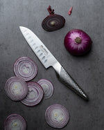Sliced red onions and a Global santoku chef's knife on a grey surface. Fluted santoku knife for effortless slicing, dicing and chopping vegetables, cutting meat, slicing fish and dicing onions.